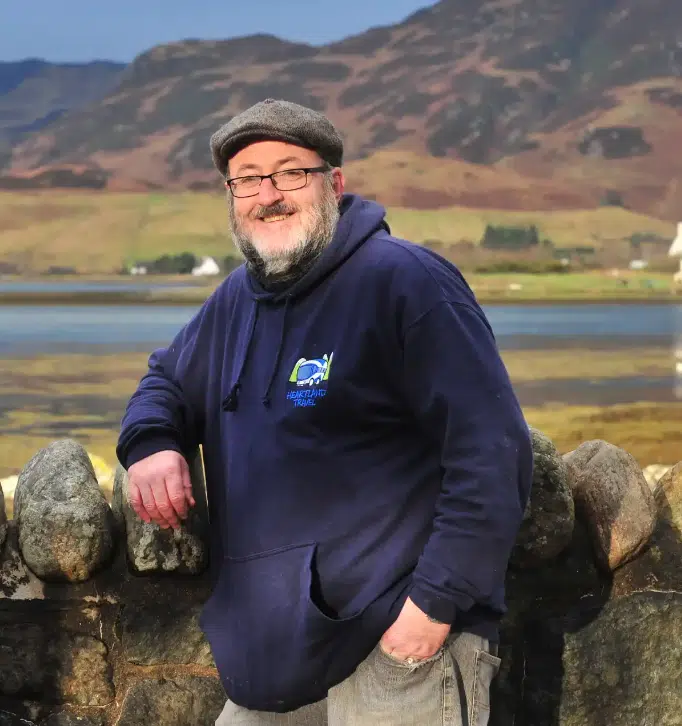Meet Our Scottish Tour Guides 1 A smiling man in a navy blue hoodie with the 'Heartland Travel' logo leans casually on a stone wall. He is wearing a flat cap, glasses, and has a short beard. Behind him is a scenic view of a calm body of water, grassy shoreline, and rolling hills under a partly cloudy sky